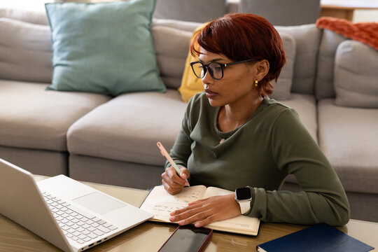 African American woman wearing olive top writing in notebook at coffee table with laptop