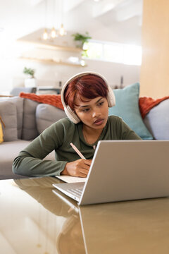 African woman leaning, watching laptop, writing in notebook at home with white headset olive top
