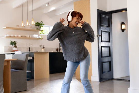 Adult woman dancing and singing in home kitchen while wearing white headphones and gray sweater