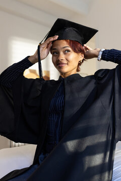 African American woman adjusting mortarboard cap and gown while sitting on bed near window blinds