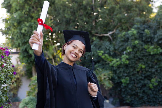 African American woman smiling, holding diploma with red ribbon in garden wearing gown, mortarboard