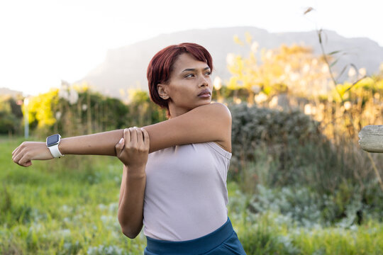 African American woman in 20s stretching right arm across chest in meadow wearing white smartwatch