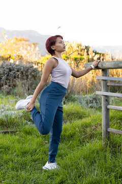 Female performing quad stretch in grassy meadow near wooden fence, wearing tank top and smartwatch