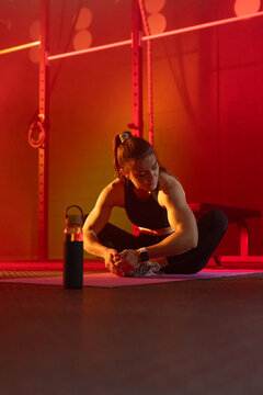 Female gym-goer stretching on purple mat in gym wearing black sportswear, squat rack, water bottle