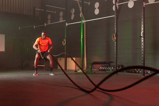 African man using battle ropes in gym, gripping ropes with kettlebells and rig visible, copy space