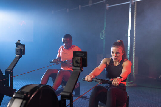 Diverse training partners rowing on ergometers in dim gym, wearing athletic clothes and smartwatch