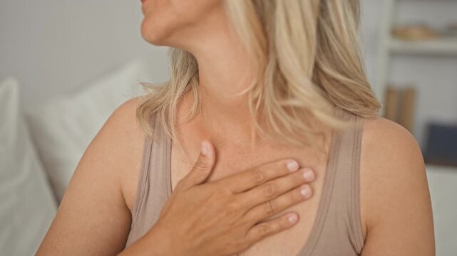 Woman, blonde middle age, hand on chest touching upper sternum and neckline, seated on bed in a building interior; serenity.