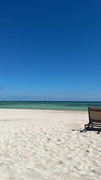 Vertical Video of beautiful white sand beach and blue waters in Cancun Mexico. Camera pans across the beach from palm tree to empty beach chairs.