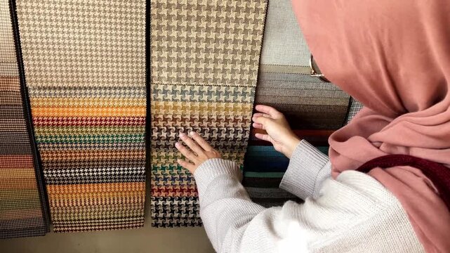 Young Asian Muslim woman in hijab examining and choosing fabric swatches and color samples for interior design project in a shop.