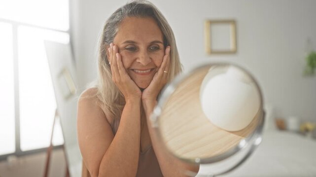Woman touching cheeks in front of a round mirror, hands and cheek visible, smiling in a bright bedroom studio with bed and decor visible; self care serenity.