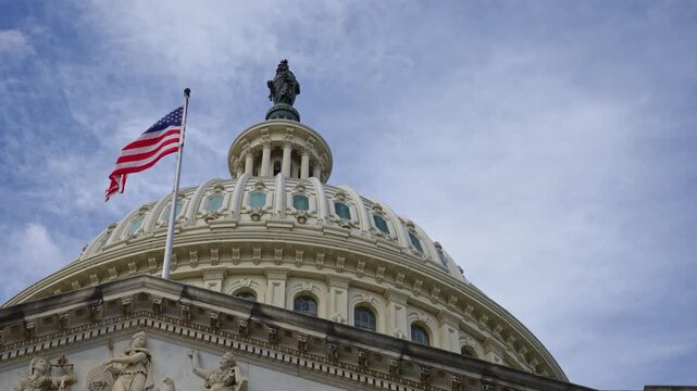 American flag waving on Capitol. American flag in clouds over US government building. American flag representing freedom of Amerika. American flag on historic Capitol dome in Washington DC.