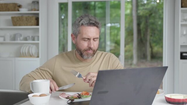 Man having lunch indoors while working on laptop in kitchen. Morning kitchen scene focused on laptop, man, and eating. Cozy interior kitchen routine with man eating and laptop work.