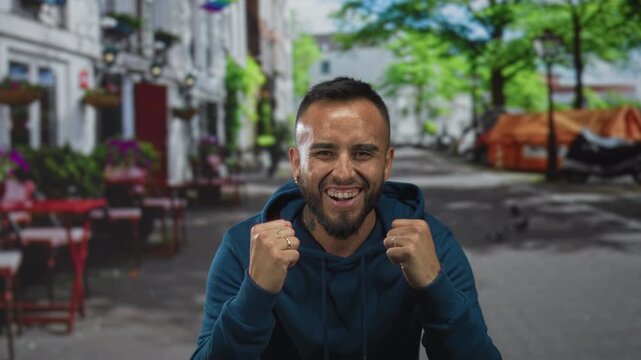 Young hispanic man in blue hoodie clenching fists and smiling broadly while standing at a restaurant terrace on a street lined with tables and trees; joy celebration success.