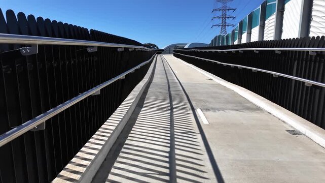 Walking perspective along concrete ramp of the elevated Federation Trail pedestrian overpass in Yarraville, with vertical screening palisades, stainless steel handrails. Melbourne, Victoria, Australia