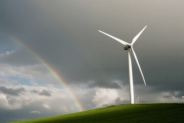 Obraz premium Wind turbine on grassy hill with rainbow against stormy sky