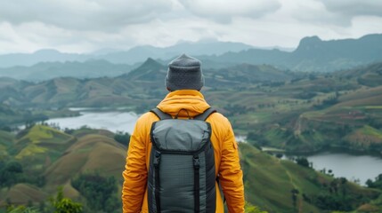 Fototapeta premium View from behind of a person wearing a yellow jacket and backpack standing on a hill overlooking a wide green mountainous terrain with lakes under a cloudy sky