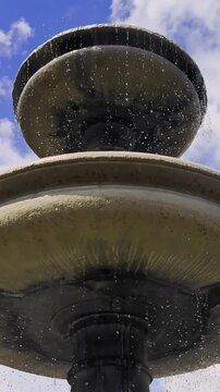 Low-angle vertical view of Turjak Fountain on Novi trg in Ljubljana, Slovenia, with water cascading over stacked stone tiers against a bright blue sky.