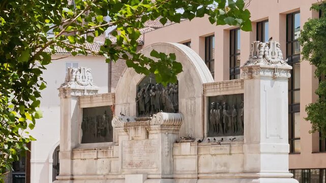 Frontal view of the Ara Giuseppe Verdi memorial in Parma, Italy, with bronze narrative reliefs, monumental stonework and sculpted figures framed by leafy trees.