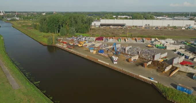 Aerial view of a heavy industrial crane grabbing scrap metal and transferring recyclable metal waste into a truck beside a waterway at a recycling yard.