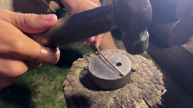 Close-up shot of hands using a hammer to forge copper wire on a small anvil outdoors at night. Ideal for artisan crafts, DIY tutorials, and metalworking videos.