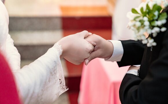Close-up of a bride and groom holding hands during a wedding ceremony with soft blurred background elements.