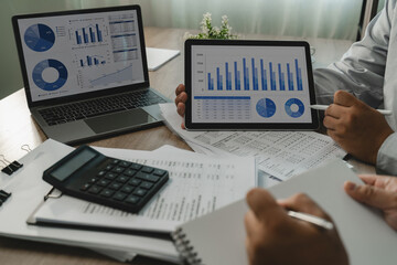 Business team reviews charts on a laptop and tablet beside printed reports and calculator,...