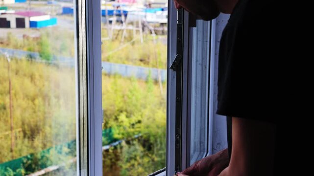 A man in a black t-shirt adjusts a window latch with a small tool, pausing to inspect the mechanism before securing it. The background reveals greenery and distant structures through the glass.