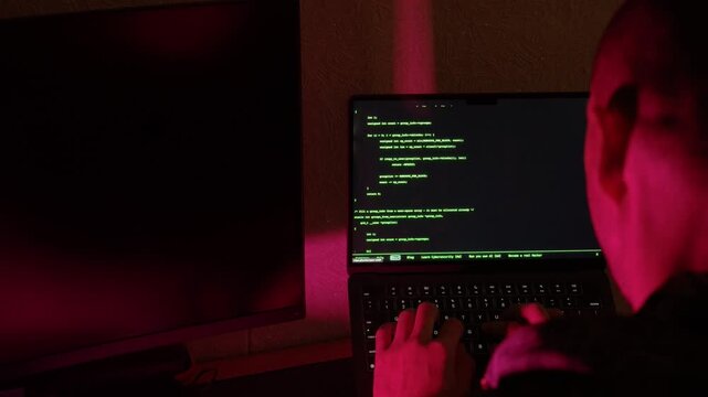 A person with a shaved head types on a laptop in a dimly lit room as ambient lighting shifts through blue, purple, and red hues. The screen displays green code that briefly highlights a text block
