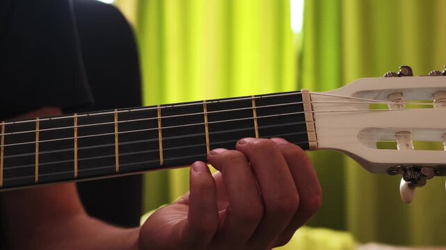 A hand forms and transitions between chords on a classical guitar fretboard against a blurred green curtain backdrop.