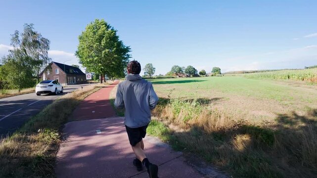tracking shot of a young man in sportswear and jacket jogging down a red asphalt bicycle path through a rural countryside landscape under a clear blue sky.