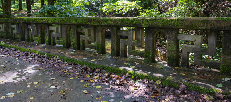 A mossy stone bridge railing decorated with Buddhist manji patterns spans across a forest stream near a Korean temple entrance.