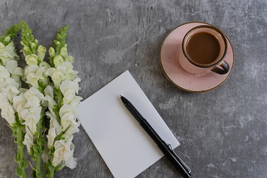 White snapdragons sit beside a pen and notepad. Pink cup holds warm coffee on a matching saucer. Stillness suggests quiet thought or creative pause