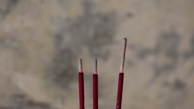 Three incense sticks stand upright. Tip of each glows softly with ember. Smoke curls gently upward, hazy and slow. Blurred stone background adds calm, meditative tone. Ideal for spiritual, healing