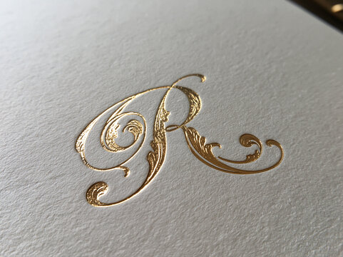 Ornate gold foil embossed calligraphy letter R monogram on textured white paper background.