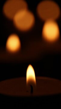 In complete darkness, a tiny spark ignites and grows into a glowing tea light candle flame, while warm orange bokeh lights gradually appear and brighten softly in the blurred background above.
