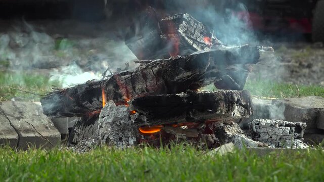 A close-up view focuses on a campfire where glowing orange embers burn deep within heavily charred and cracked wood logs.