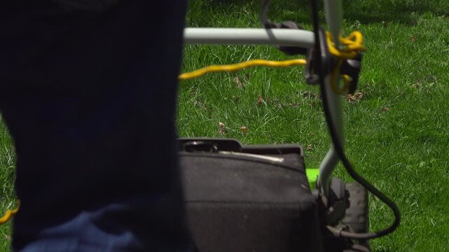A close-up shot captures a man walking into frame pushing an electric lawnmower across a green yard while holding a yellow extension cord.
