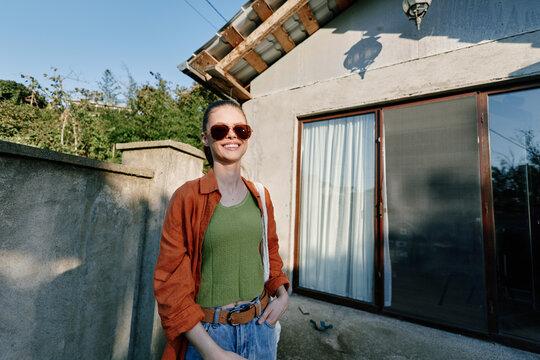 Smiling young woman in sunglasses and orange shirt stands outdoors near a rustic house wall. Casual summer style, travel lifestyle and relaxed vacation mood concept.