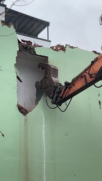 A construction site with a large excavator demolishing a green wall. Debris is scattered around, indicating ongoing demolition work.