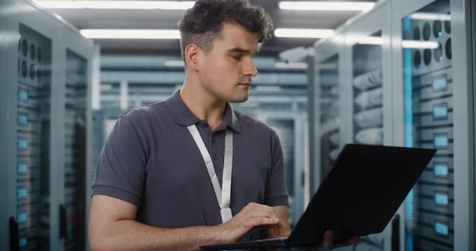 Focused Network Engineer Stands in Server Room, Using Laptop to Configure Enterprise Hardware. Cyber Security and Cloud Storage Management Protect Critical Data Systems. Infrastructure Monitoring.