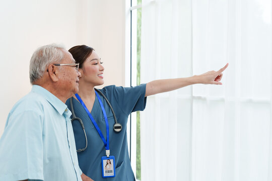 Senior Asian man standing with young female caregiver nurse in blue scrubs pointing and looking out the window in cozy living room at nursing home. Physical therapy, healthcare concept