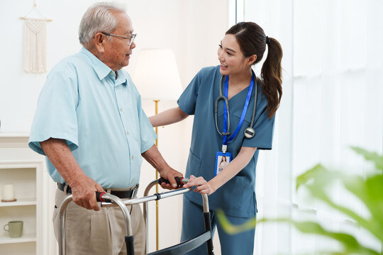 Young friendly female caregiver nurse in blue scrubs is smiling and helping Asian senior man practice walking with walker in cozy living room at nursing home. Physical therapy, healthcare concept
