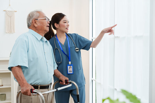 Senior Asian man standing with young female caregiver nurse in blue scrubs pointing and looking out the window in cozy living room at nursing home. Physical therapy, healthcare concept