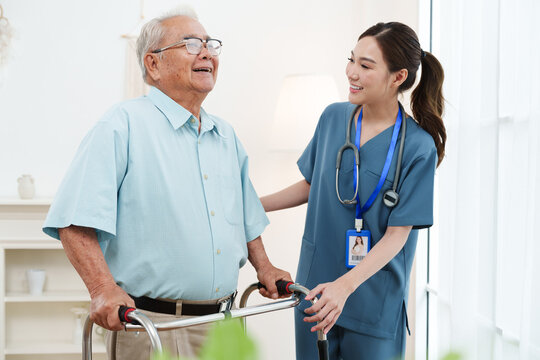 Young friendly female caregiver nurse in blue scrubs is smiling and helping Asian senior man practice walking with walker in cozy living room at nursing home. Physical therapy, healthcare concept