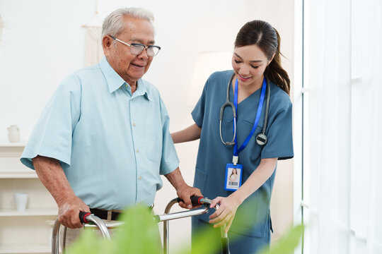 Young friendly female caregiver nurse in blue scrubs is smiling and helping Asian senior man practice walking with walker in cozy living room at nursing home. Physical therapy, healthcare concept
