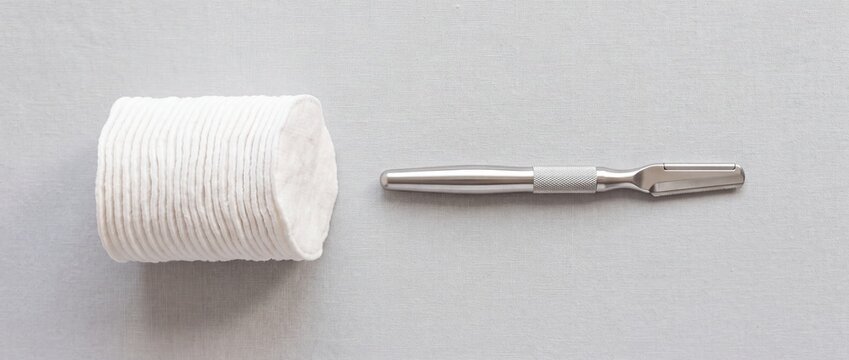Professional dermaplaning razor tool lies next to stack of white cotton pads on minimalist grey fabric surface for facial hair removal