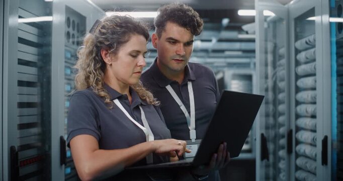 In Modern Data Center Server Room, Female IT Specialist Works on Laptop While Male Colleague Inspects Server Racks. System Administration and Teamwork Ensure Continuous Cloud Uptime. Big Data Concept.