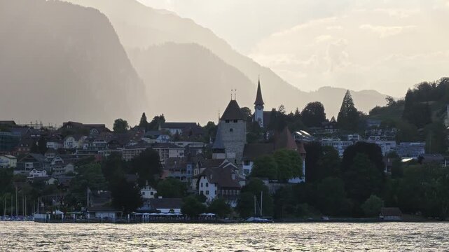 Dreamy golden hour sunset over Spiez Schiffstation (ferry terminal) and Schloss Spiez Castle on Lake Thun. 4K offshore POV from a passenger boat cruise.