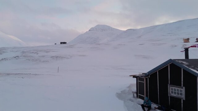 Pullback aerial reveal of a remote wooden cabin flying a Danish flag amid the Svalbard snowfield, evoking a quiet sense of isolation and shelter in the Arctic