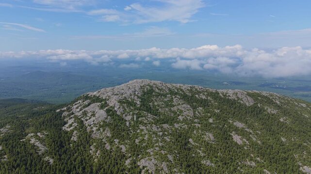 Panoramic drone shot around granite top of Mount Monadnock rises over green woodland as low clouds drift in the distance, New Hampshire, US